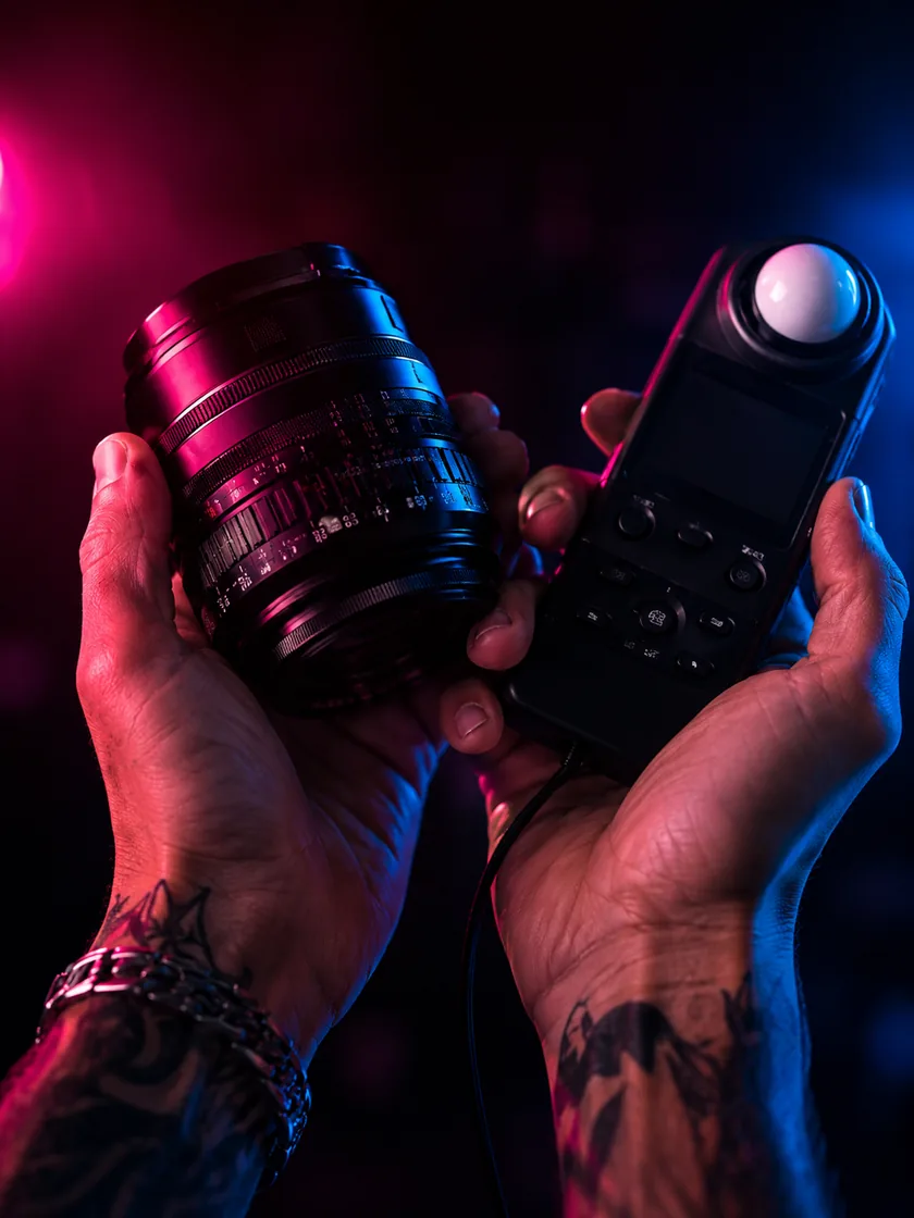 Two hands holding a prime lens and a light meter, lit cyan on one side and magenta on the other.
