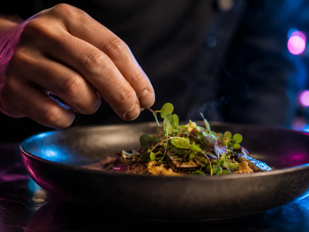 A hand placing micro herbs onto a finished bowl, close crop, steam rising.