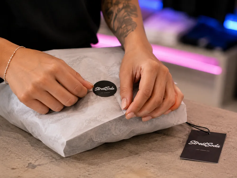 Two hands folding tissue around an order and sealing it with a branded sticker.